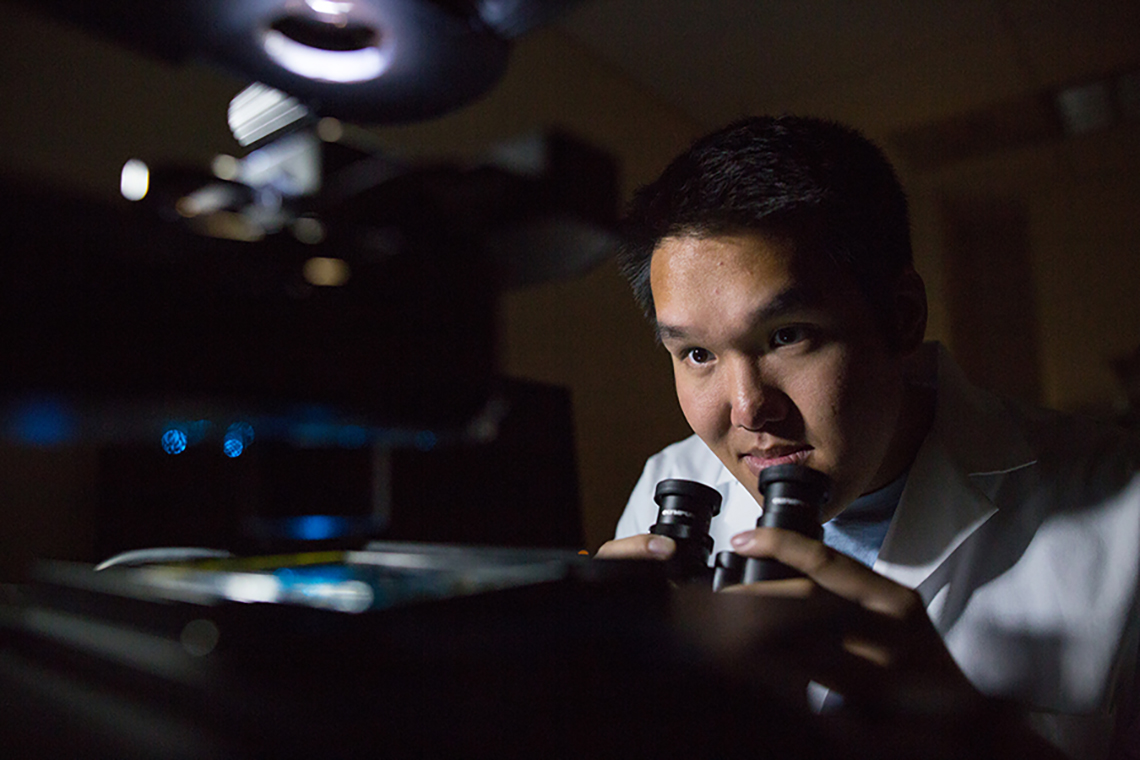 A researcher in a dark room looks up from a microscope eyepiece