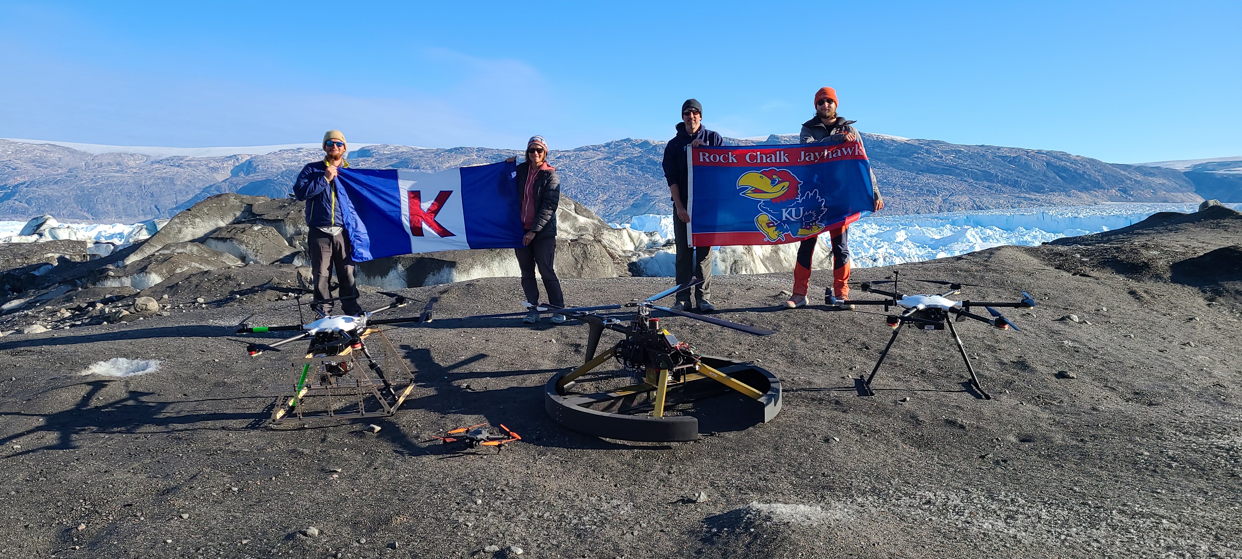 "Emily Arnold and colleagues pose near a scientific drone while holding KU flags."